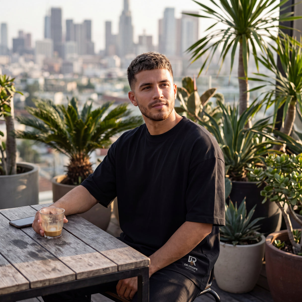 A man wearing a black t-shirt and black sweatpants sits at a wooden table on a rooftop terrace with a city skyline in the background.