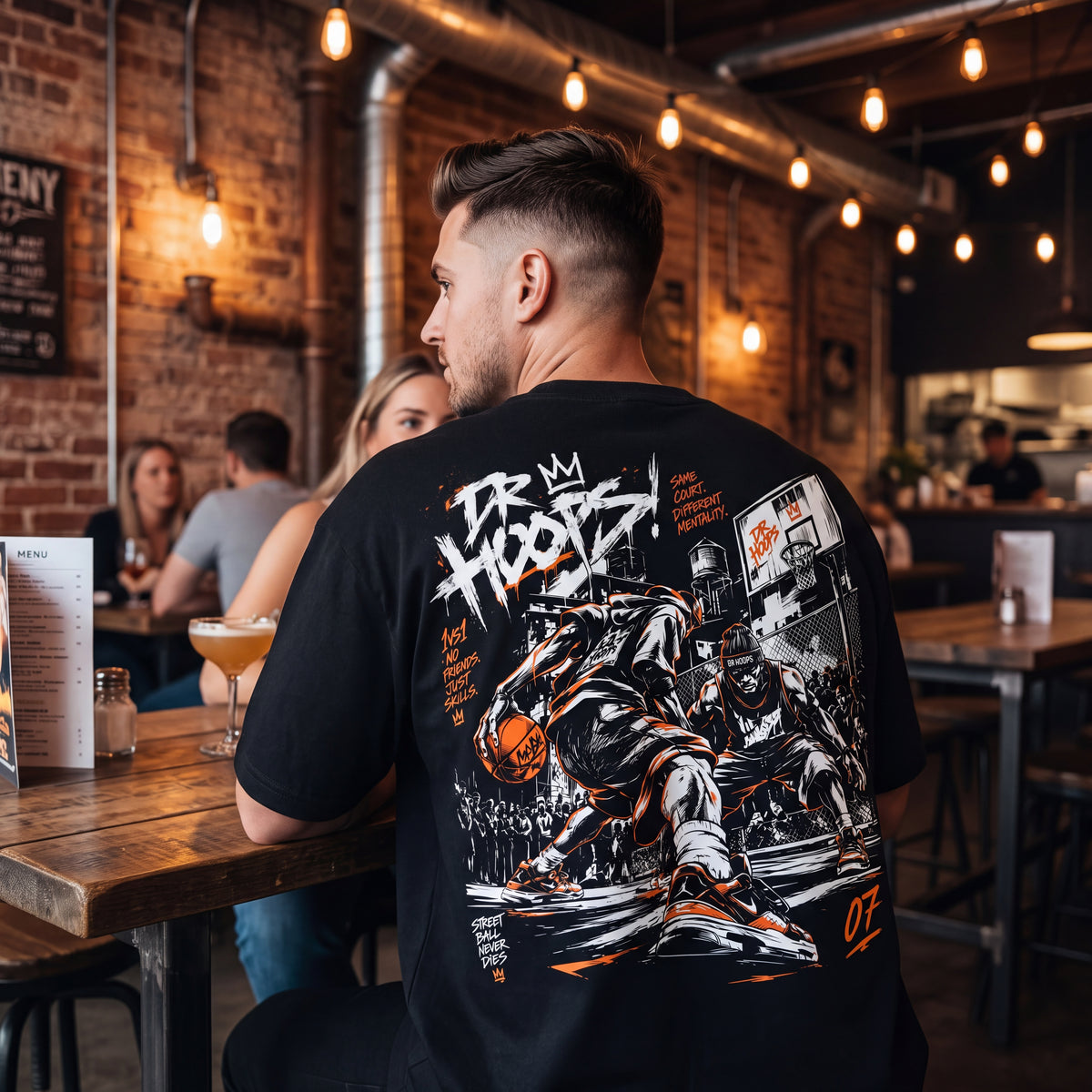 A man wearing a black graphic t-shirt with a large basketball-themed print on the back, seated at a wooden table in a warm, industrial-style restaurant.