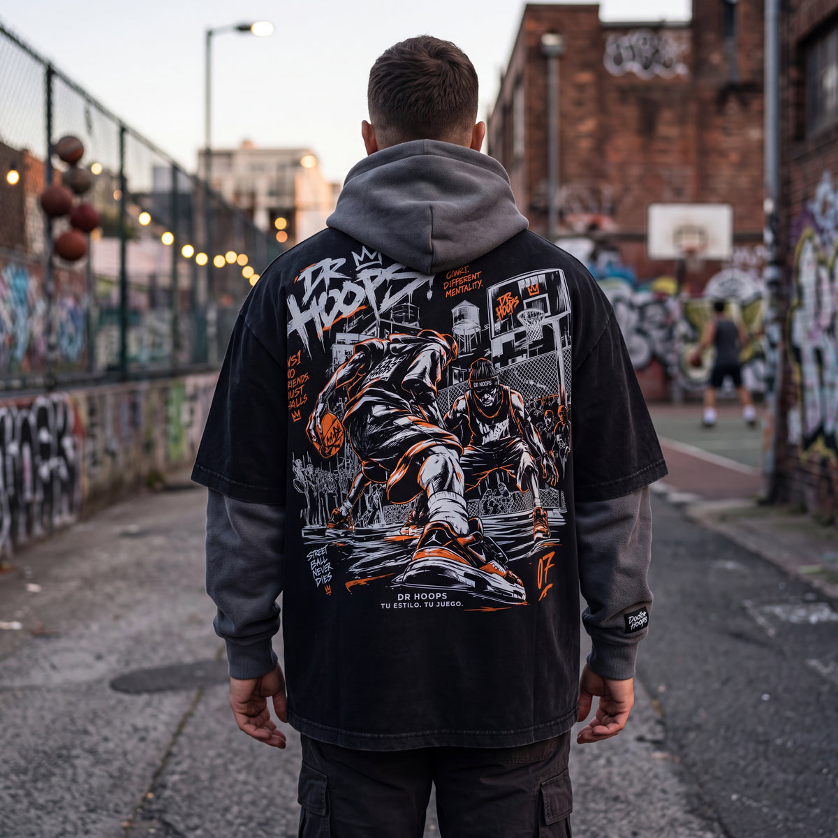 A man viewed from behind wearing a black washed graphic hoodie with a large back print, standing on an urban street with graffiti walls and a basketball court in the background.