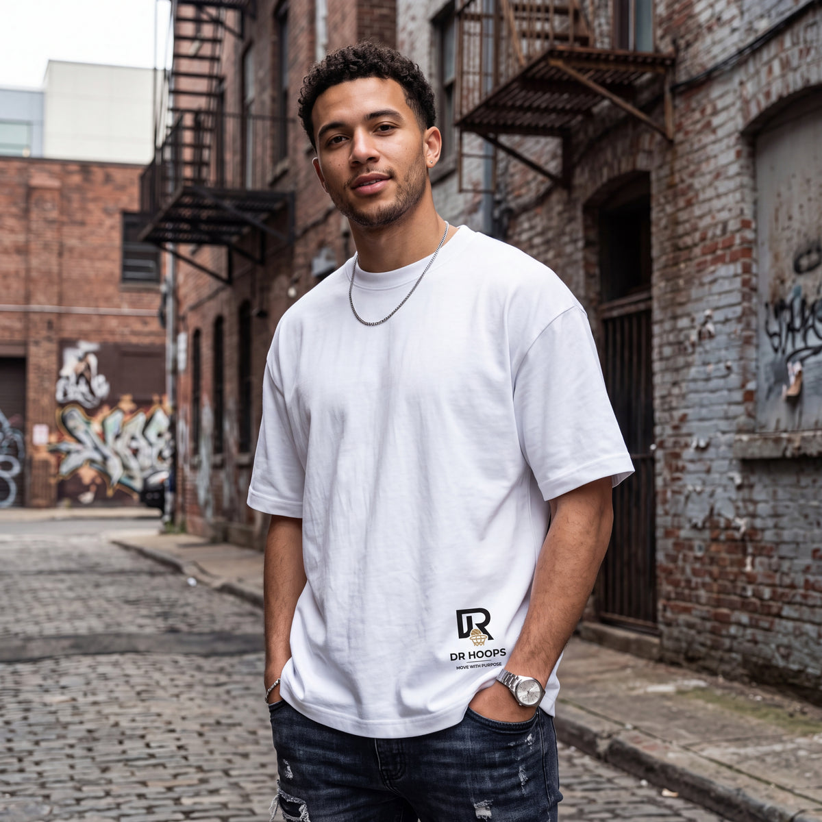 A man wearing a white crewneck t-shirt with a black 'DR HOOPS' logo on the lower left hem, standing in an urban brick alleyway.