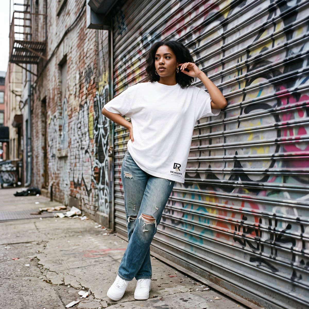 A woman posing in an urban alleyway next to a graffiti-covered metal shutter, wearing a white oversized t-shirt with a black logo, distressed blue jeans, and white sneakers.