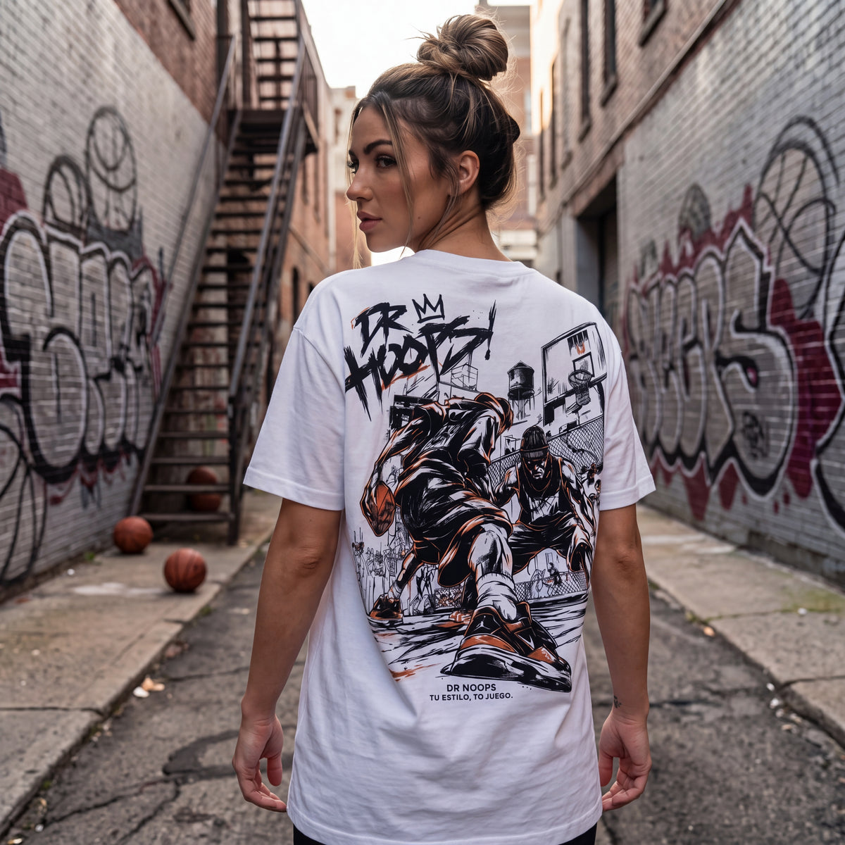 A woman stands in an urban alleyway with graffiti walls, photographed from behind to show the back of a white graphic T-shirt. The shirt features a large black and orange illustration of a basketball player with the text 'I'M HOOPS!' and smaller text reading 'I'M HOOPS TO KEEP THE HOOPS.