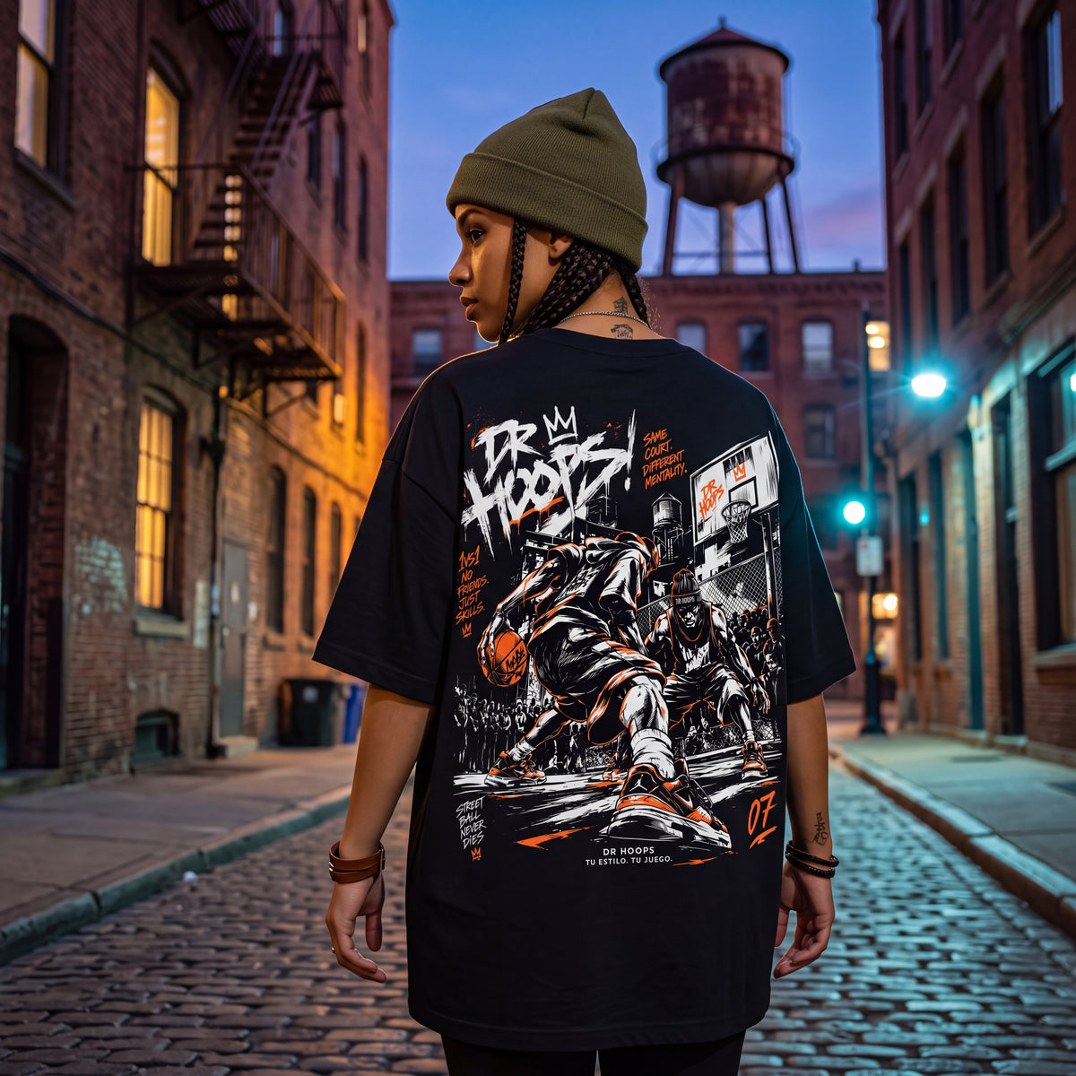 A person viewed from behind wearing an oversized black graphic T-shirt and an olive green beanie, standing on a cobblestone city street at dusk with brick buildings and a water tower in the background.